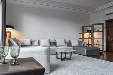 stylish large living room in white and gray tones, gray sofa, armchair, round table, wooden coffee table, carpet, floor lamp, gray curtains on windows. Illuminated by lamp. Space for relaxation