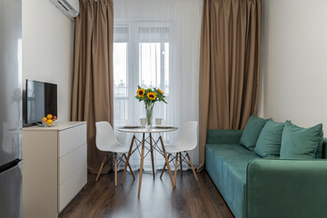 Cozy modern living room with turquoise sofa, chest of drawers, TV, round table, chairs. vase of flowers on table, yellow sunflowers in room. beige curtains on window. Space for relaxation