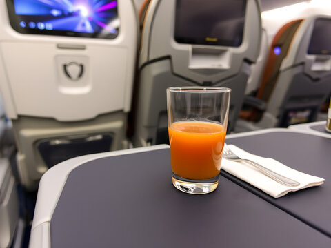 A glass of orange juice on a meal tray in economy class. The airplane seats and screens in the background highlight the sleek and practical in-flight experience. - Powered by Adobe