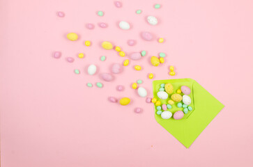 Empty white card in envelope and  scattered chocolate Easter colored candy eggs on pink background.