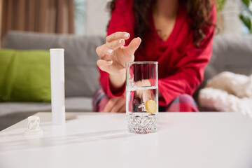 Woman in red pajama sitting on sofa in living room, drops tablet into glass of water, creating...