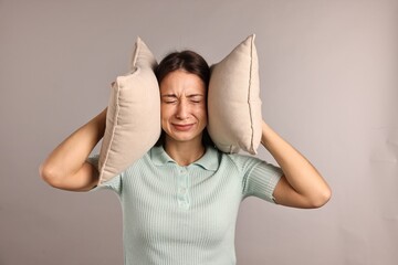 Annoyed woman covering her ears with pillows due to loud sound on light grey background