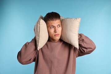 Annoyed young man covering his ears with pillows due to loud sound on light blue background