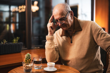 Anxious senior man with cup of coffee sitting alone in the cafe at evening. He is worried about something and having a headache.