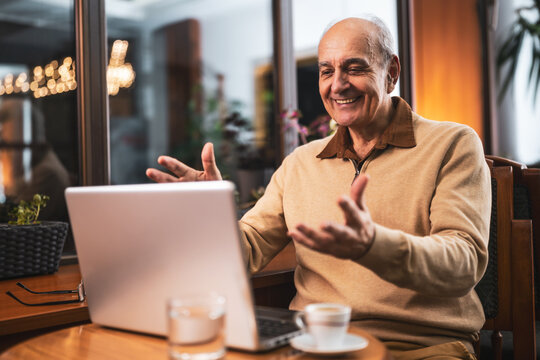 Joyful senior man in casual clothing talking and gesturing while having a video call on his laptop. He is using computer and enjoying drinking coffee and relaxing in the city cafe.