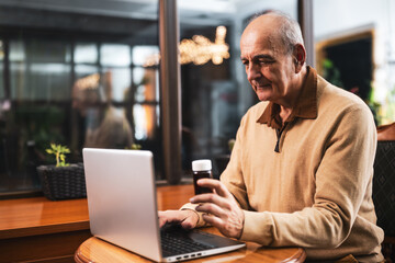 Senior man holding pill bottle and using laptop for searching more information about his prescription medicine while sitting in the cafe.	