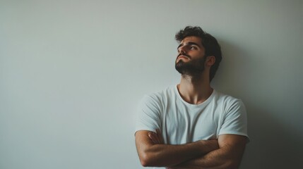 17.Man in casual attire, standing against a plain white wall with arms crossed and a thoughtful expression; subtle lighting captures a contemplative, introspective moment