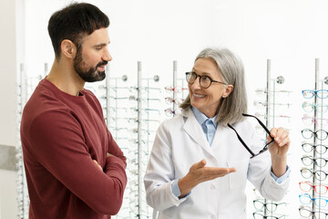 Senior female optician showing eyeglasses to customer in optical store