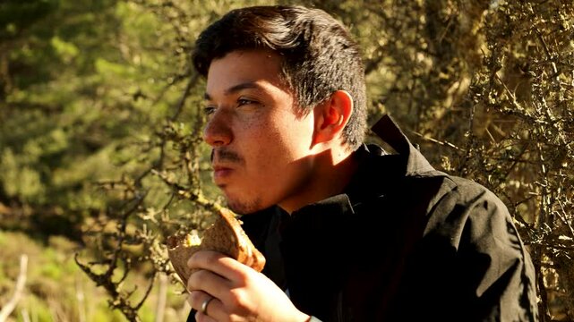 Freckled adventurous young man sitting on a mountain eating a sandwich while resting and looking around