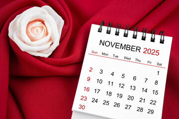 November 2025 desk calendar and pink rose on red textile.