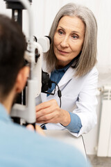 Senior female doctor performing eye exam on patient using phoropter in ophthalmology clinic