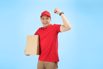 happy Asian courier fists his arm while holding a package against a blue background and wearing a red company uniform.