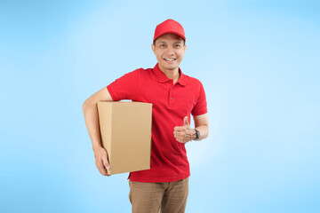 An happy Asian courier gift a thumb while holding a package against a blue background wearing red company uniform.