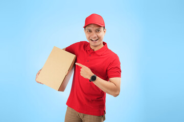 An Asian courier pointing his finger while holding a package against a blue background wearing red company uniform.