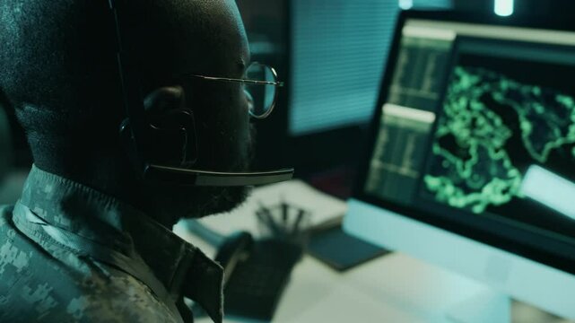 Closeup over-shoulder shot of African American male officer in camouflage uniform, with headset working at night in command center, watching satellite surveillance and giving directions to army