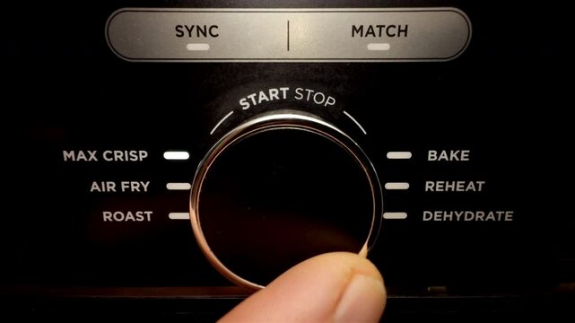 Macro closeup of a finger turning the heat setting knob of an air fryer cooker to the required cooking program option, then pressing the start button.
