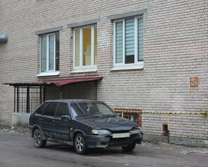 An old rusty black car is parked near a gray brick wall of a house, Iskrovsky Prospekt, Saint...
