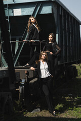 three girls dressed in black suits. They are on the train platform next to the freight car.