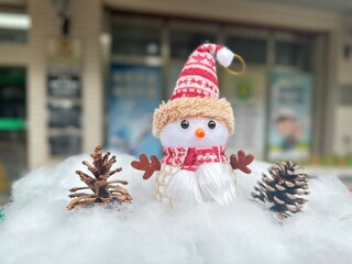 An adorable snowman doll wearing a red and white knitted hat and scarf, decorated with pinecones on...