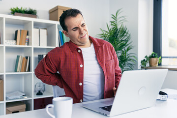 Tired business man with back pain looking uncomfortable while working with laptop in a modern...