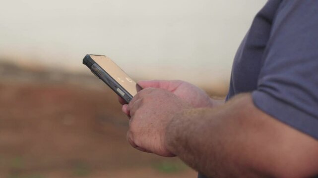 Close up shot of Caucasian man hand typing and tapping on smartphone while standing in field outdoors. Male fingers texting on cellphone in countryside on sunny day. Technology concept

