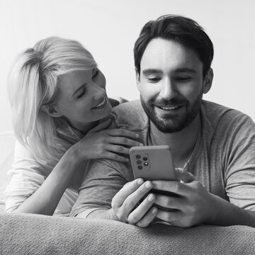 Monochrome bw image - couple lying on bed browsing internet reading text message use cellphone cell phone cellular device. Happy bearded man and woman shop online by smartphone. Technology. Square