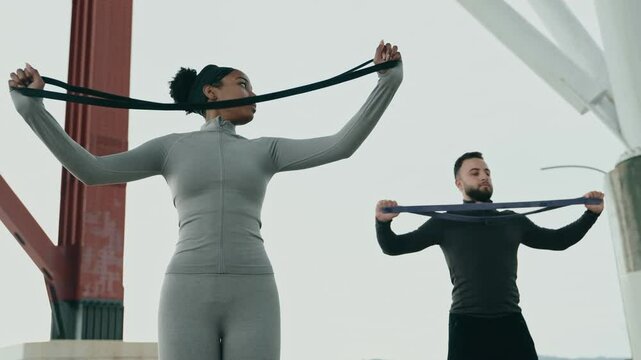 Two people engaged in resistance training with elastic bands under an open sky, focusing on strength and fitness outdoors.