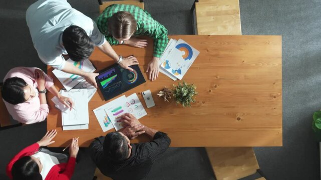 Top view of business man using tablet display increasing graph while diverse marketing people sharing idea or planing strategy at meeting table with stock market statistic scatter around. Convocation.