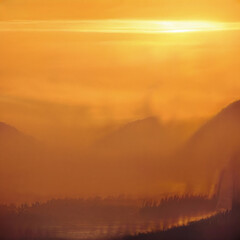 Obraz premium A warm, orange-toned landscape featuring silhouetted mountains and trees under a bright, hazy sky, possibly at sunrise or sunset.