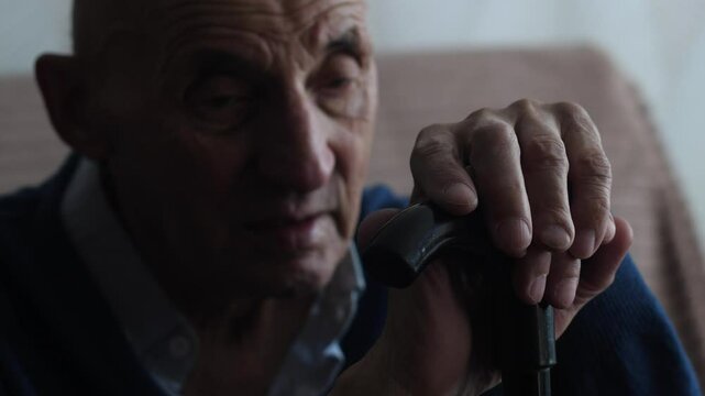 elderly caucasian man 70-79 years old sits thoughtfully holding his hands on a walking stick, pensioner sits alone with a walking stick while inside
