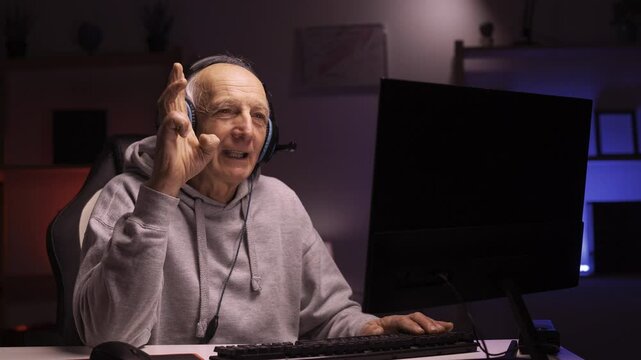 Senior mature gamer streaming his nighttime gaming session using personal computer counting fingers on camera at home gaming room