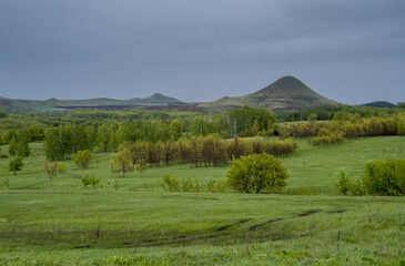 A valley covered with trees and bushes against a backdrop of low mountains. The sun breaks through the clouds.