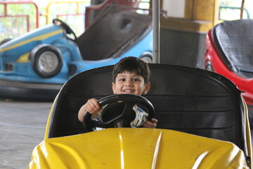 cute little 4 year old boy is enjoying the dashing car ride in the amusement park. He is driving...