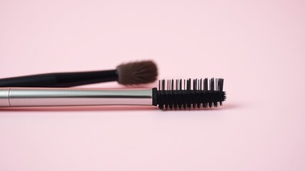 A close up of a black mascara wand with a silver handle and black bristles on a pink background, a makeup brush lies behind it
