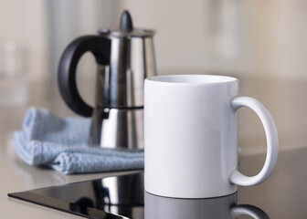 White coffee mug on induction stove against moka pot on blue dish cloth closeup, mockup