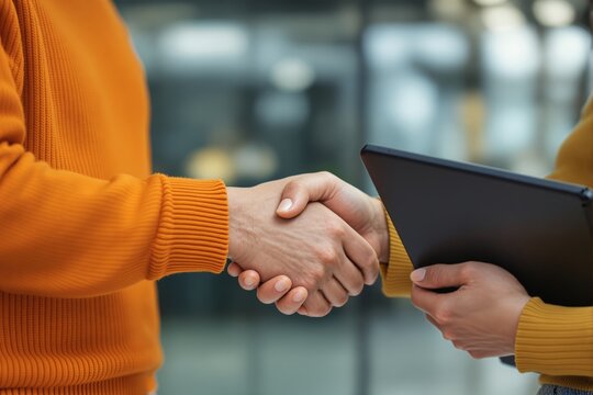 Close-up handshake of two individuals in casual orange sweaters symbolizing business agreement or partnership indoors.