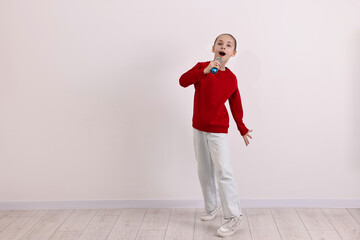 Little girl with microphone singing near white wall. Space for text