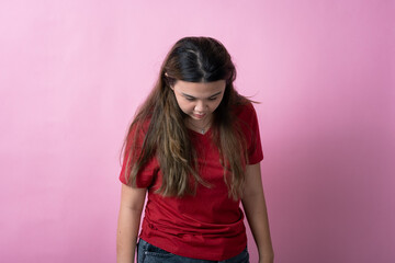 Asian woman in a red shirt with long brown hair stands while looking down with a neutral or somber expression, arms relaxed at her sides, against a solid pink background.