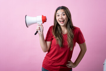 Young woman with long brown hair wearing a red t-shirt, smiling and holding a white and red megaphone against a solid pink background. She poses confidently, looking directly at the camera.