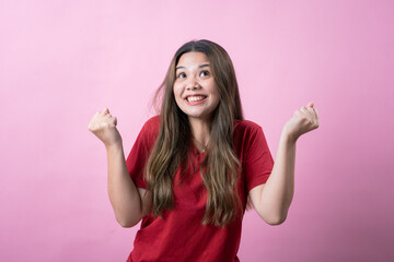 Young woman in casual red t-shirt with long brown hair, standing against a solid pink background, expressing excitement and happiness with clenched fists and wide smile, energetic positive vibe.