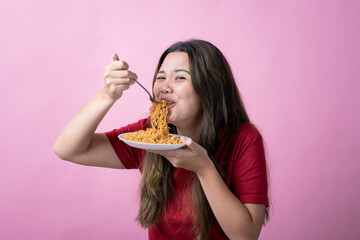 Asian woman in a red shirt enjoys eating noodles from a white plate using a fork, smiling playfully and looking at the camera against a solid pink background.