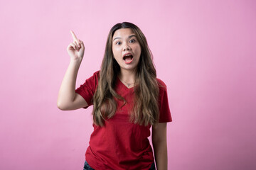 Asian woman in a red t-shirt and jeans standing against a pink background, smiling brightly with excitement, raising one finger upward as if having a new idea or solution