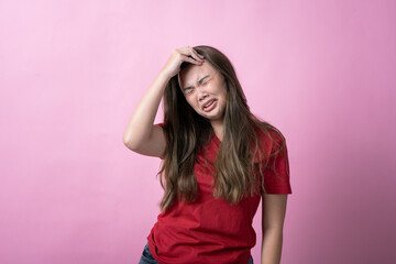 Asian woman in a red shirt with long brown hair grimaces in frustration, holding her head with one hand and grabbing her elbow with the other, standing against a solid pink background.