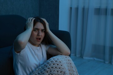 Fear of darkness. Scared young woman with blanket near couch indoors at night, space for text