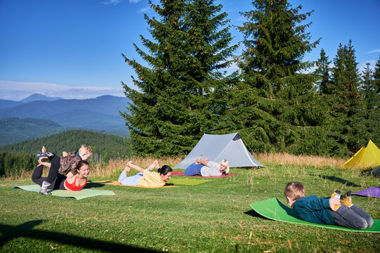 Group of people doing yoga pose outdoor in camping in the mountains. Adults and children on yoga mats, each doing a yoga pose under a clear blue sky in the morning. Young boy is instructor.