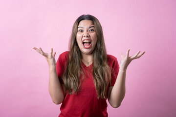 Frustrated young woman in a red shirt makes a confused gesture with both hands, showing irritation against a pink background. The playful and exaggerated facial expression adds humor and personality.