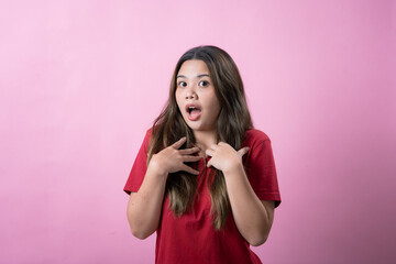 A surprised young woman with long brown hair and light skin, wearing a red T-shirt, places her hands on her chest and opens her mouth in shock, standing against a solid pink background.