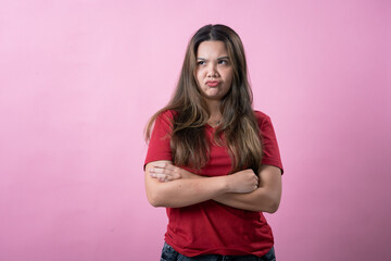 Asian woman in a red shirt with long brown hair crosses her arms and makes a pouty, dissatisfied expression against a vibrant pink background.