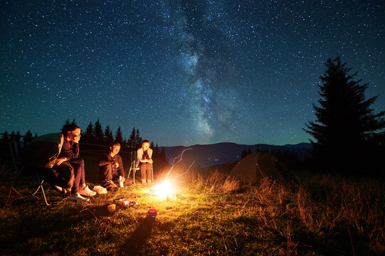 Group of friends sits around campfire under magnificent starry sky, with Milky Way above. Tents pitched nearby. Serene mountain landscape creates perfect setting for night of stargazing, camaraderie.