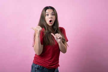 Excited Asian woman in red shirt points to herself and gestures behind her against a vibrant pink background.